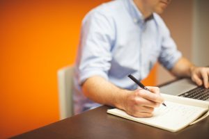 Man writing in a notebook beside a laptop against an orange wall