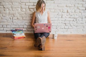 Woman sitting on the floor against a brick wall working on a laptop beside a stack of books
