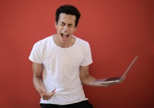 Man shouting in frustration while holding a laptop against a red background