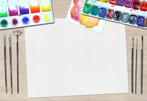 Blank sheet of paper surrounded by watercolour paint palettes and brushes
