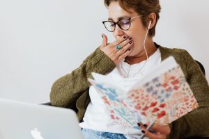 Woman in glasses yawning while holding an illustrated book at a laptop